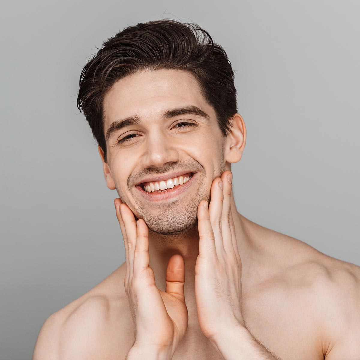 Beauty portrait of half naked smiling young man touching his face and looking at camera isolated over gray background