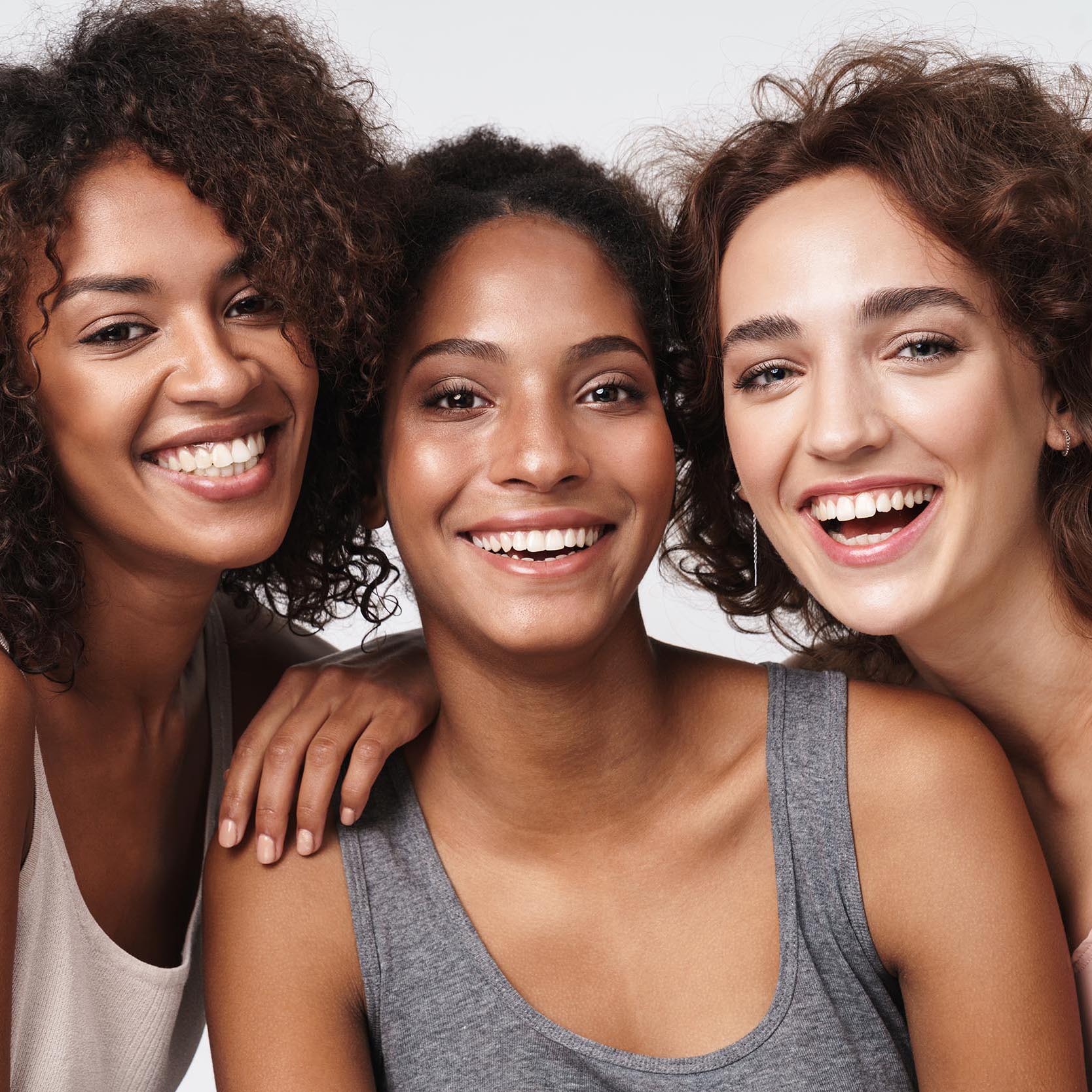 Portrait of three young multiracial women standing together and smiling at camera isolated over white background